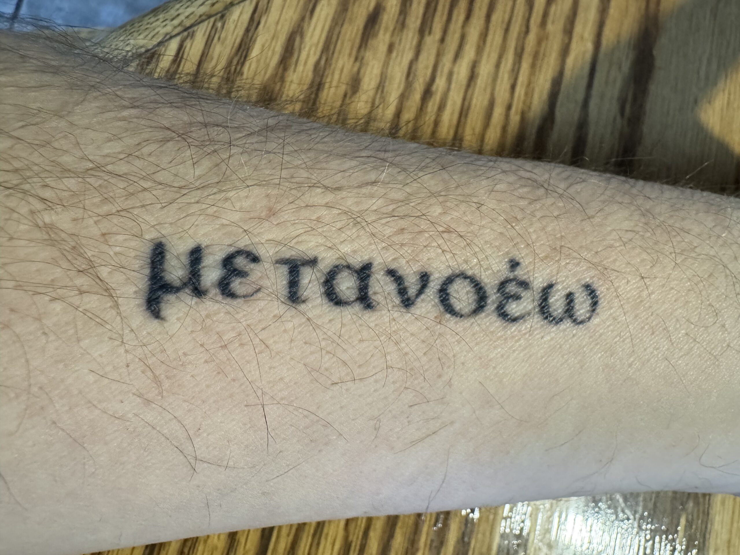 Black ink tattoo of the Greek word metanoia in elegant script lettering, symbolizing spiritual transformation and change of thinking.